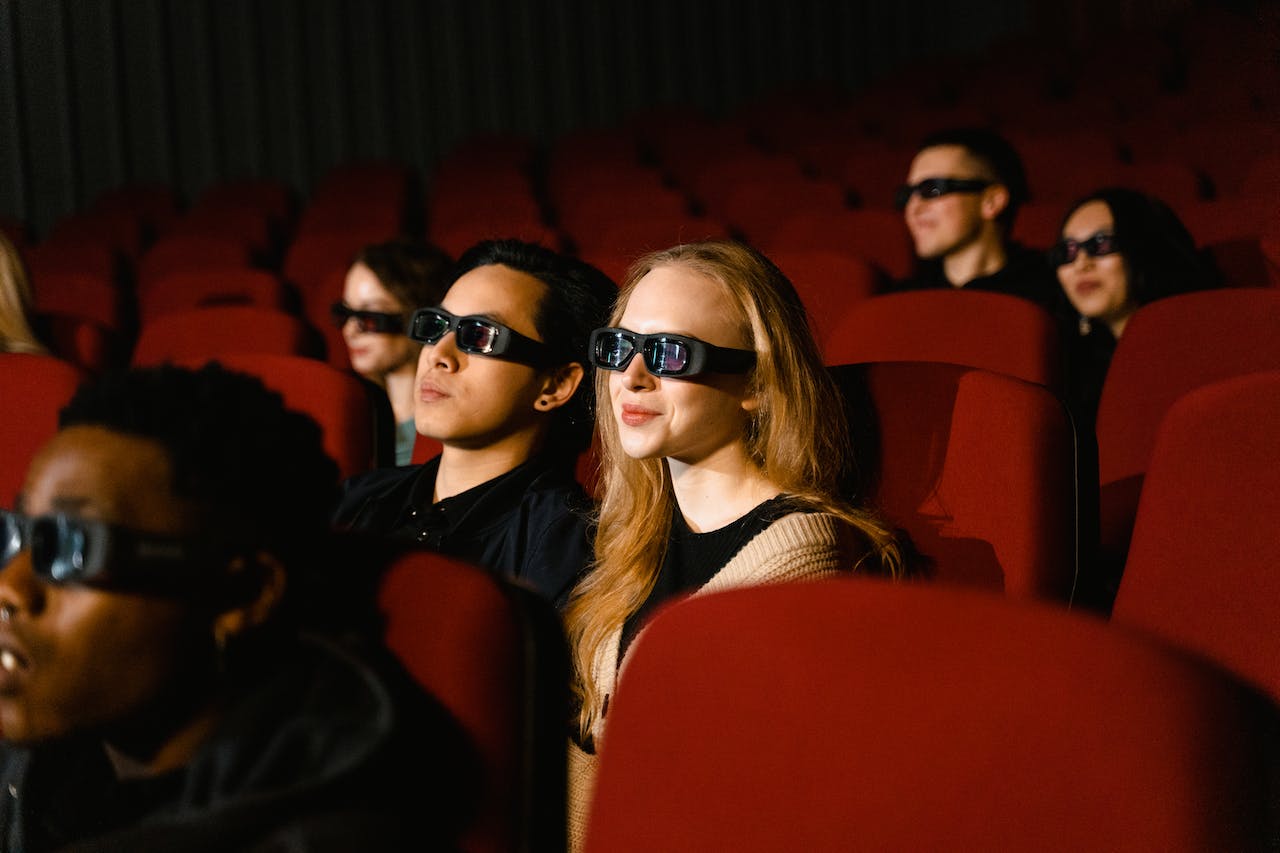 Kuvassa näkyy joukko ihmisiä istumassa elokuvateatterissa, jännittyneinä ja innostuneina katselemassa 3D-laseilla varustettua elokuvaa. Heidän kasvoillaan heijastuu jännitys ja odotus, kun elokuvamaailma herää eloon heidän edessään